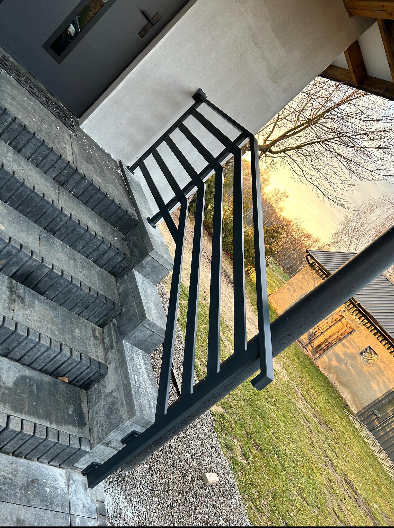 Nowoczesna, czarna balustrada schodowa z geometrycznym wzorem, zamontowana na zewnątrz budynku. Widok z góry, schody z szarej kostki, trawnik w tle.