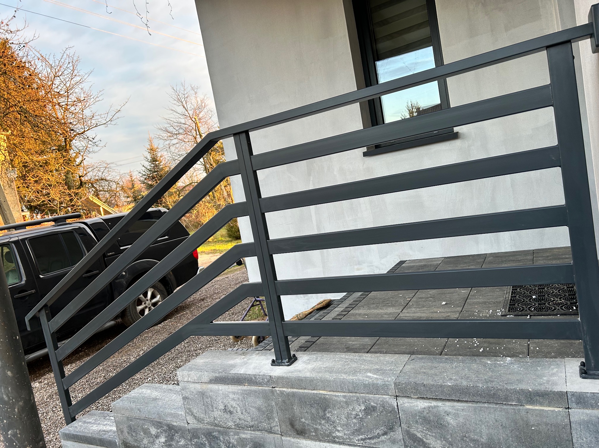 Nowoczesna, czarna balustrada metalowa z poziomymi szczeblami, zamontowana przy schodach wejściowych do domu. Widoczny fragment samochodu i otoczenie domu.