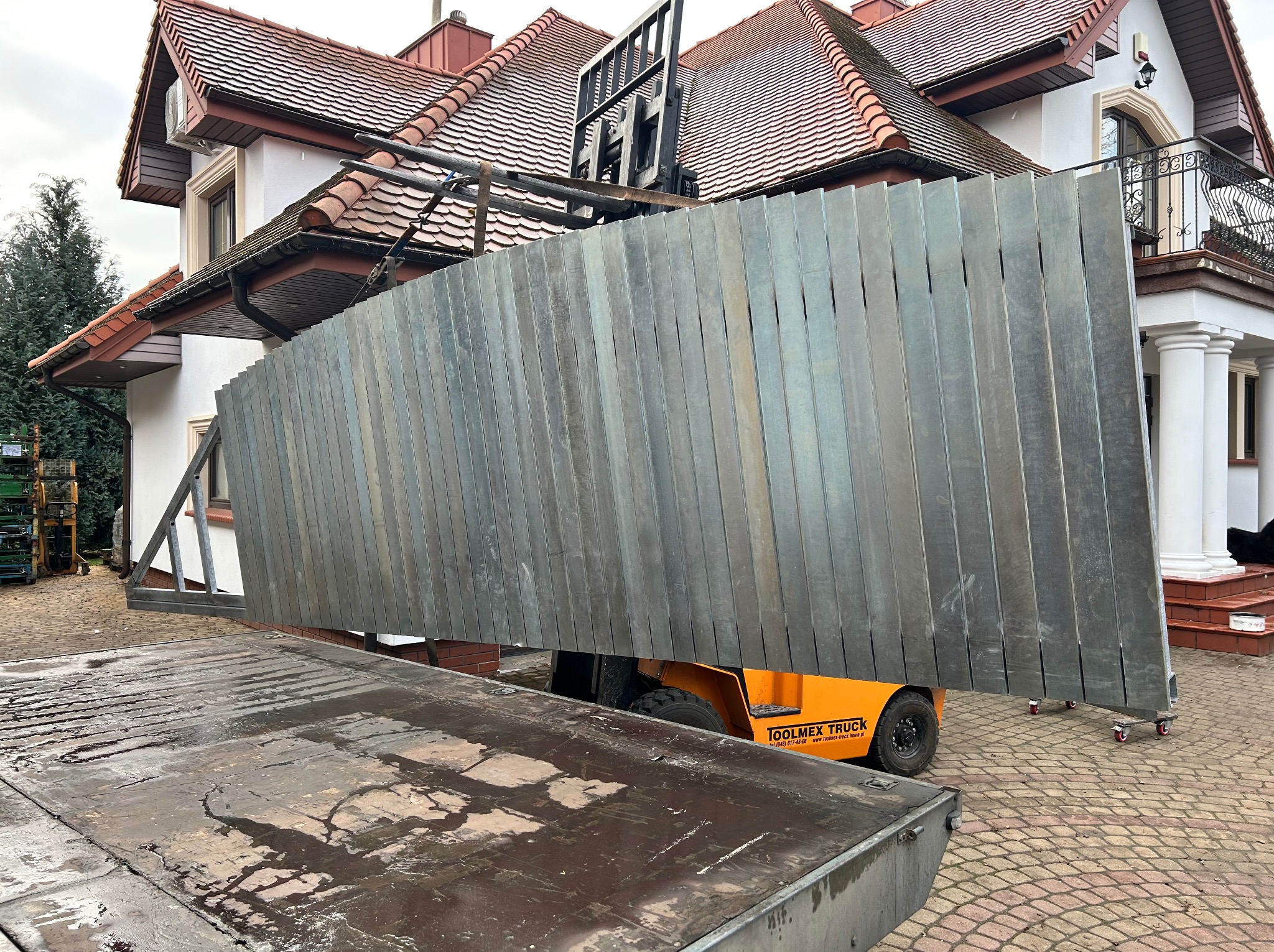 Transport elementów metalowych na budowę domu z balkonem. Widoczny wózek widłowy i fragment ciężarówki. Elementy w kolorze srebrnym, ustawione pionowo.