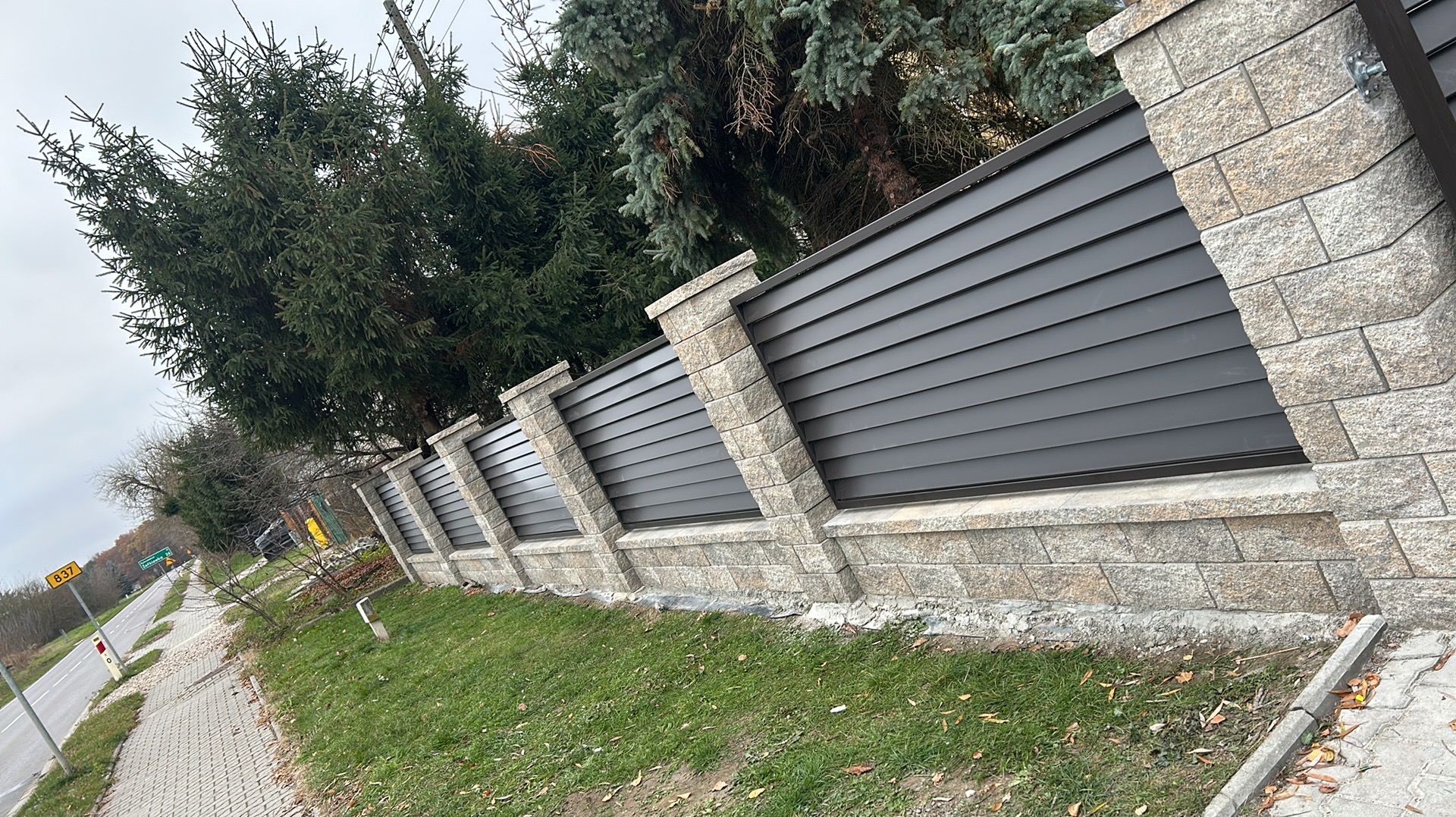 Nowoczesne ogrodzenie z szarymi, poziomymi panelami i kamiennymi słupkami, widoczne z poziomu chodnika, z drzewami w tle. Element posesji przy drodze.