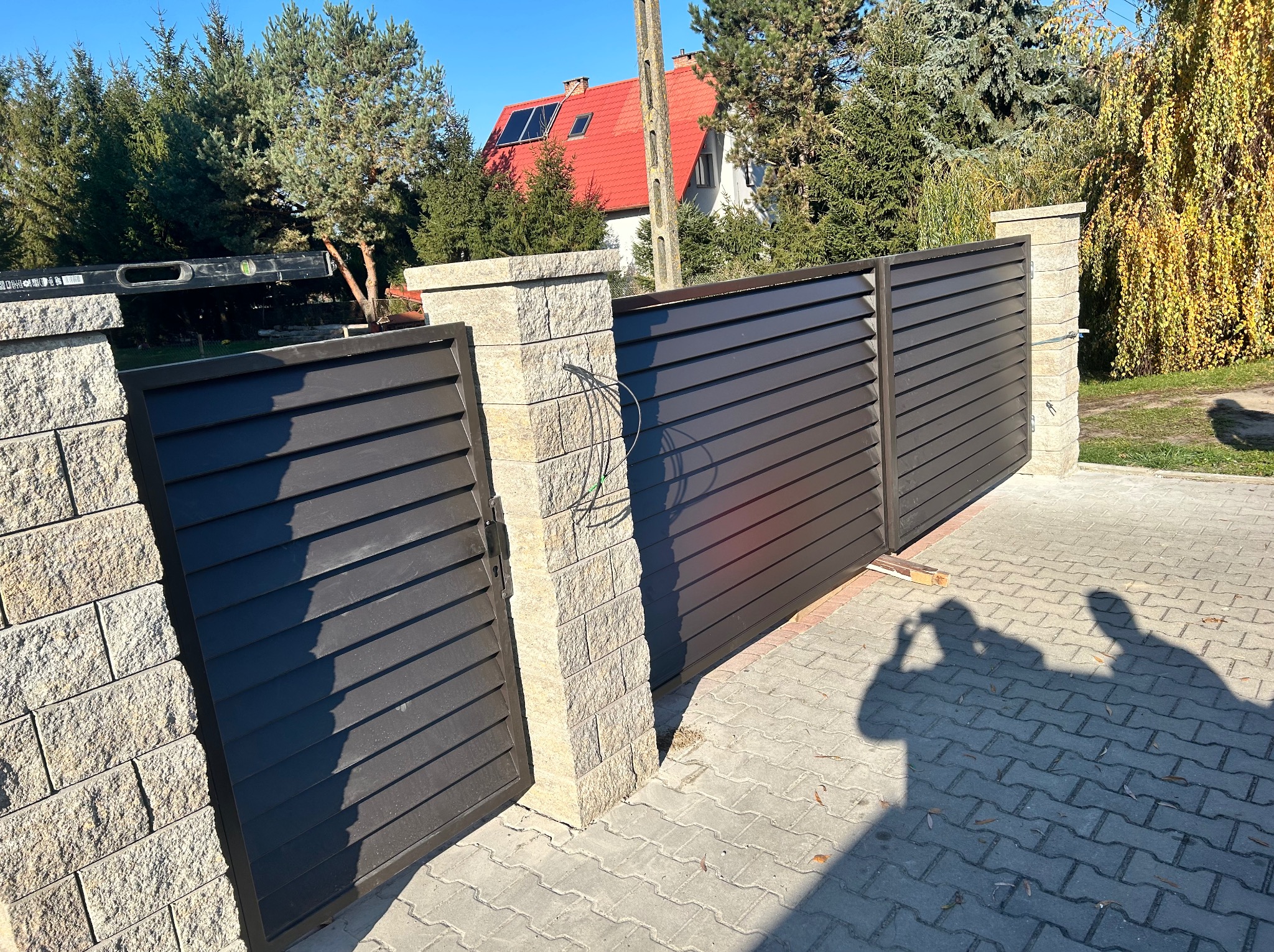 Nowoczesna brama posesyjna z poziomych paneli w kolorze antracytu, zamontowana na słupkach z jasnego kamienia łupanego. Widoczne okablowanie.