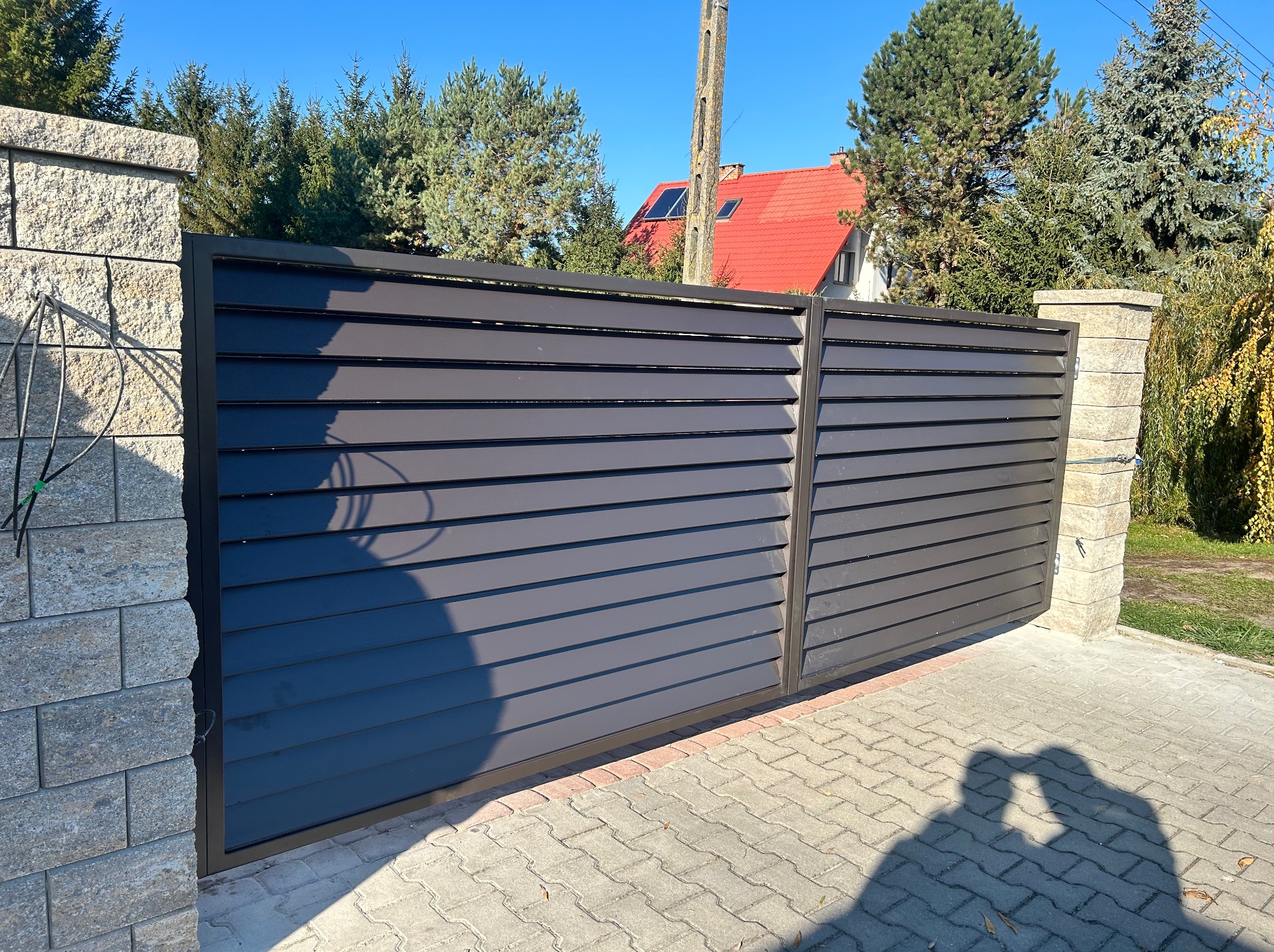 Nowoczesna, przesuwna brama w kolorze antracytu z poziomymi panelami, zamontowana na kamiennych słupkach. W tle widoczny dom z czerwoną dachówką.