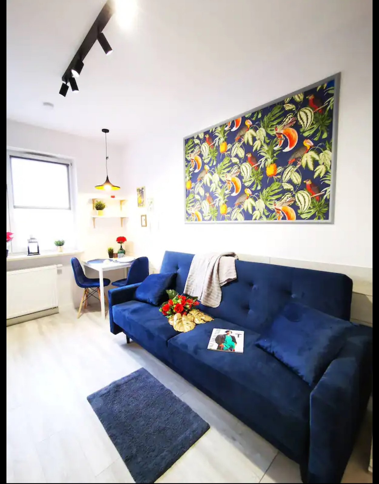 Wykończone wnętrze pokoju dziennego: niebieska sofa z poduszkami, wzorzysty obraz na ścianie, stół z krzesłami w tle, dywanik na podłodze. Nowoczesne oświetlenie.