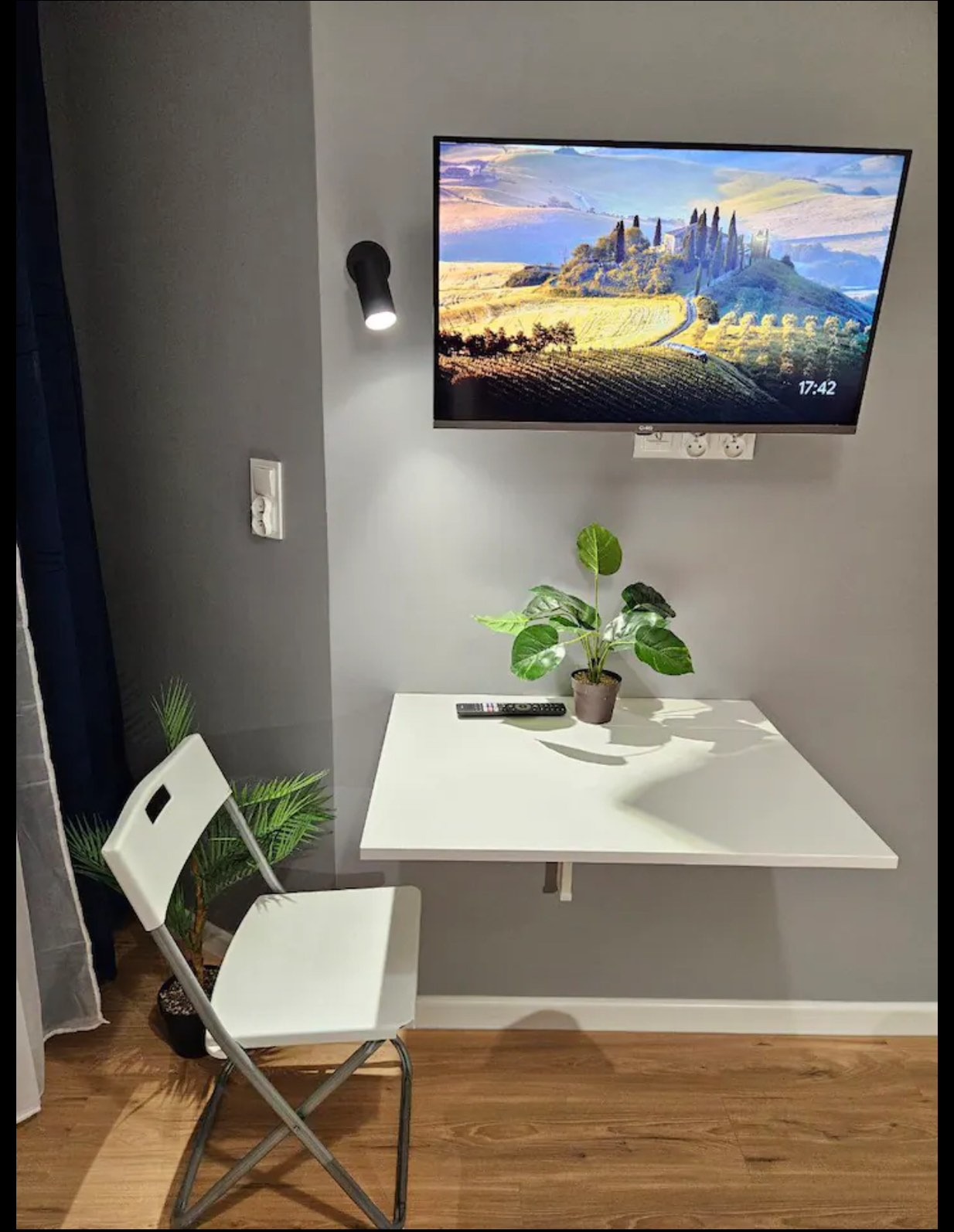 Szara ściana z telewizorem i składanym blatem, na którym stoi roślina. Obok krzesło, a na ścianie lampa i gniazdko. Widoczna podłoga z paneli.