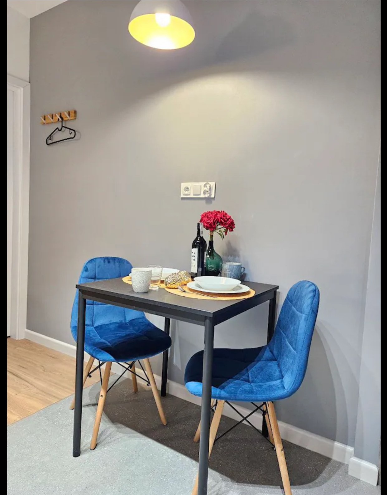 Stół z dwoma krzesłami w pokoju z szarą ścianą. Na stole wazon z kwiatami, butelka wina i zastawa stołowa. Nad stołem wisi lampa.