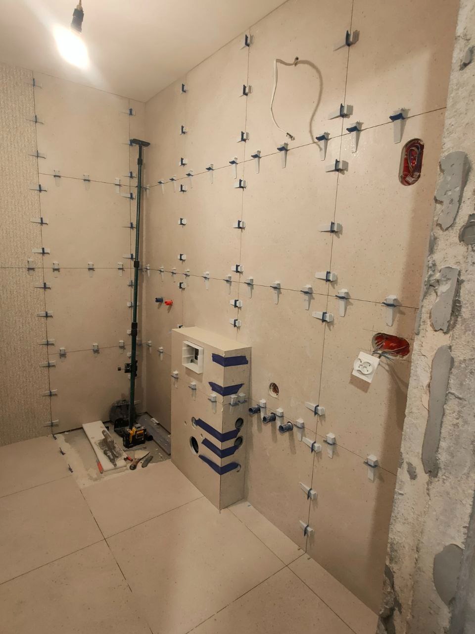 Układanie płytek w łazience, widoczne krzyżyki dystansowe, surowy stan ściany po prawej. Etap prac przed fugowaniem. Jasne płytki podłogowe i ścienne.