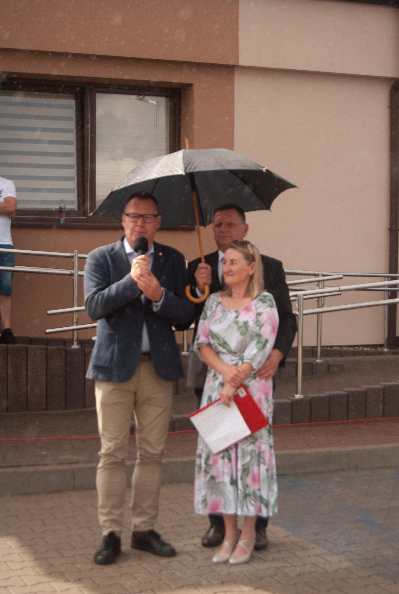 Uroczystość na zewnątrz w deszczu: mężczyzna w garniturze przemawia do mikrofonu pod parasolem trzymanym przez drugiego mężczyznę, obok stoi kobieta z dokumentami.