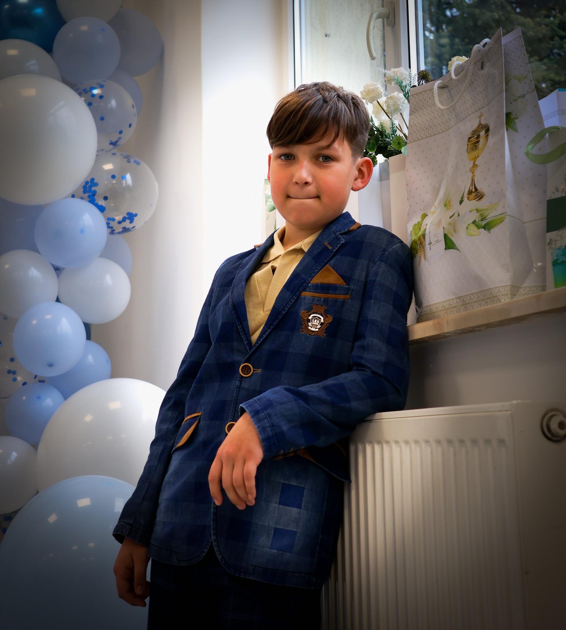Chłopiec w niebieskiej marynarce oparty o grzejnik, obok dekoracje z białych i błękitnych balonów oraz torby prezentowe. Portret w jasnym wnętrzu.
