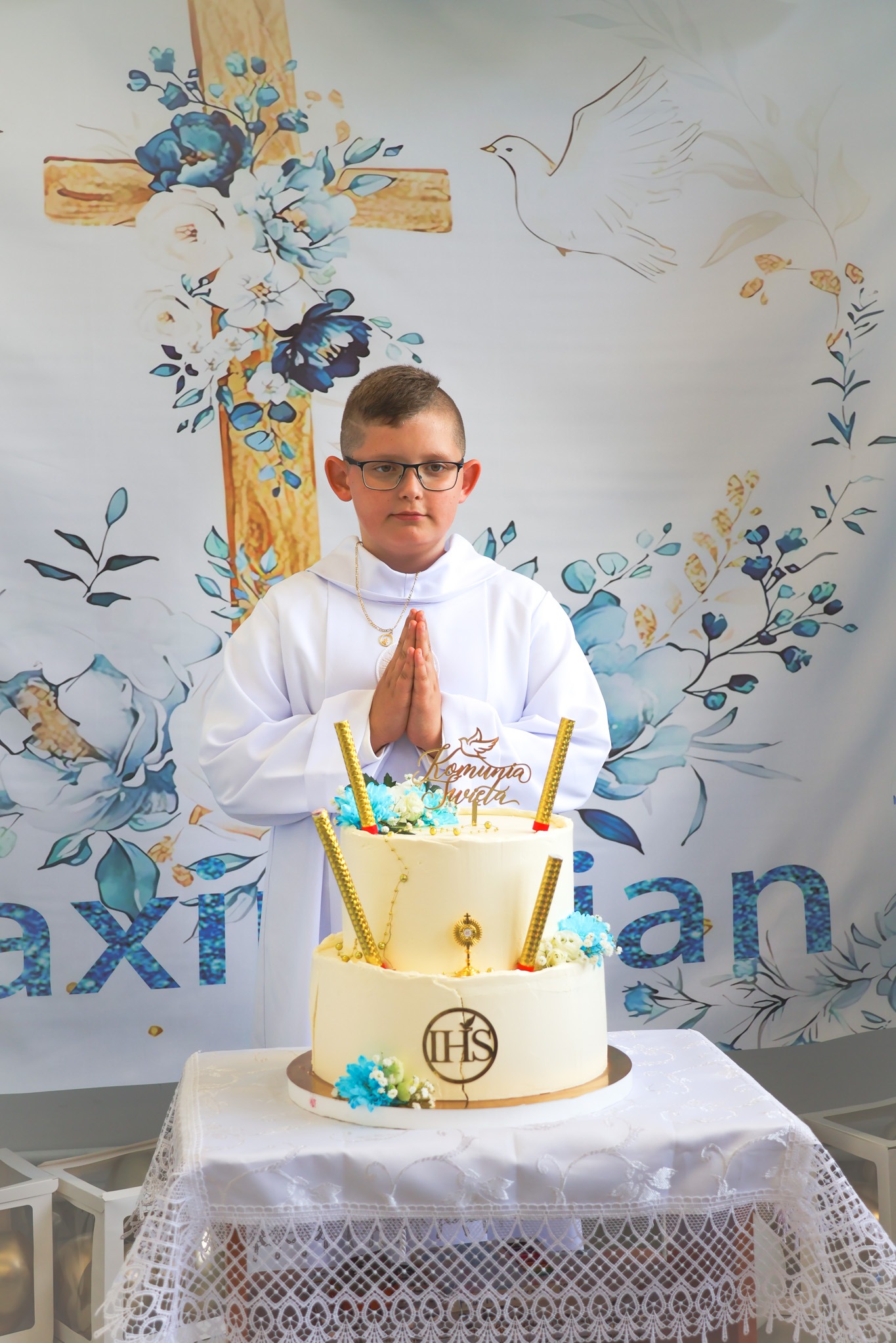 Chłopiec w albie komunijnej, ze złożonymi rękami, stoi za dwupoziomowym tortem komunijnym na tle dekoracji z krzyżem i gołębiem. Ujęcie centralne, bliskie.