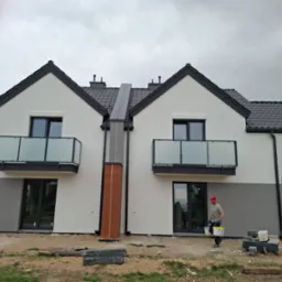 Nowoczesny, dwukondygnacyjny dom bliźniak z balkonami i szarą elewacją w dolnej części. Pracownik w czerwonej czapce przenosi wiadra z farbą.