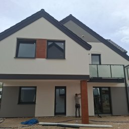 Serhii Kupriianchuk - Dwukondygnacyjny dom z elewacją w odcieniach bieli i szarości, z elementami dekoracyjnymi imitującymi drewno. Pracownik wykonuje prace wykończeniowe przy kolumnie.