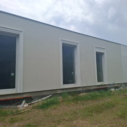 Serhii Kupriianchuk - Jasna elewacja budynku z trzema oknami o białych ramach, widok z zewnątrz, trawa i elementy instalacji przy fundamencie. Pochmurne niebo w tle.