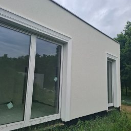 Serhii Kupriianchuk - Boczny widok domu z jasną elewacją, dużymi oknami z białymi ramami i zielonym otoczeniem. Widoczna faktura tynku i detale wykończenia okien.