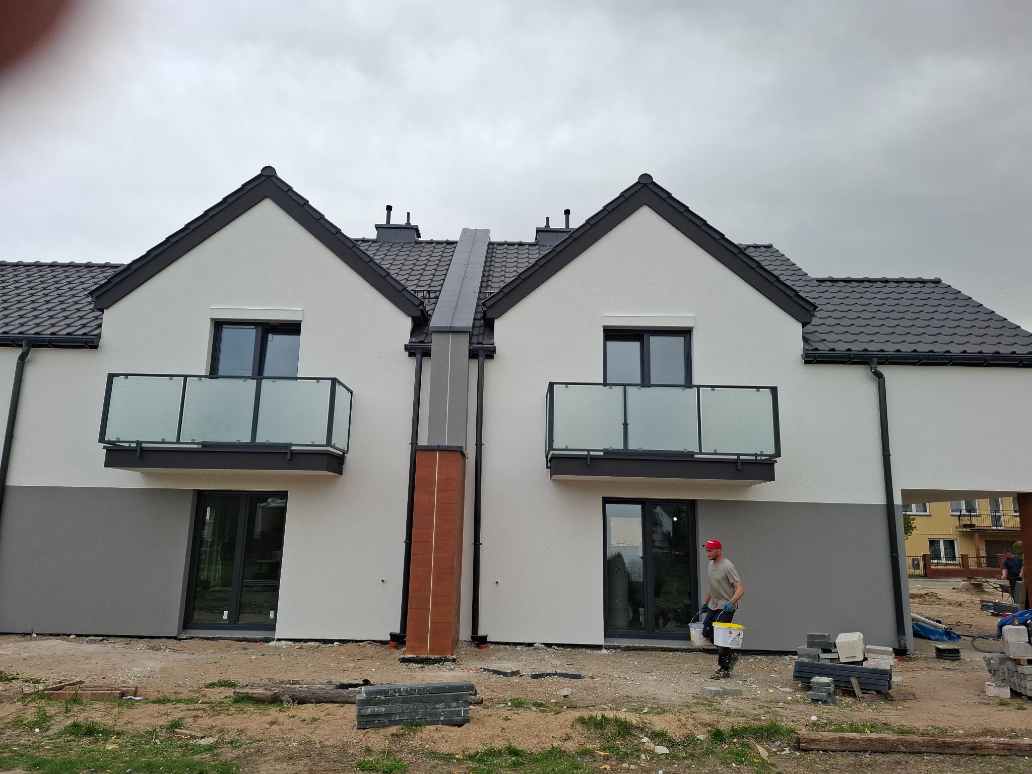 Nowoczesny, dwukondygnacyjny dom bliźniak z balkonami i szarą elewacją w dolnej części. Pracownik w czerwonej czapce przenosi wiadra z farbą.