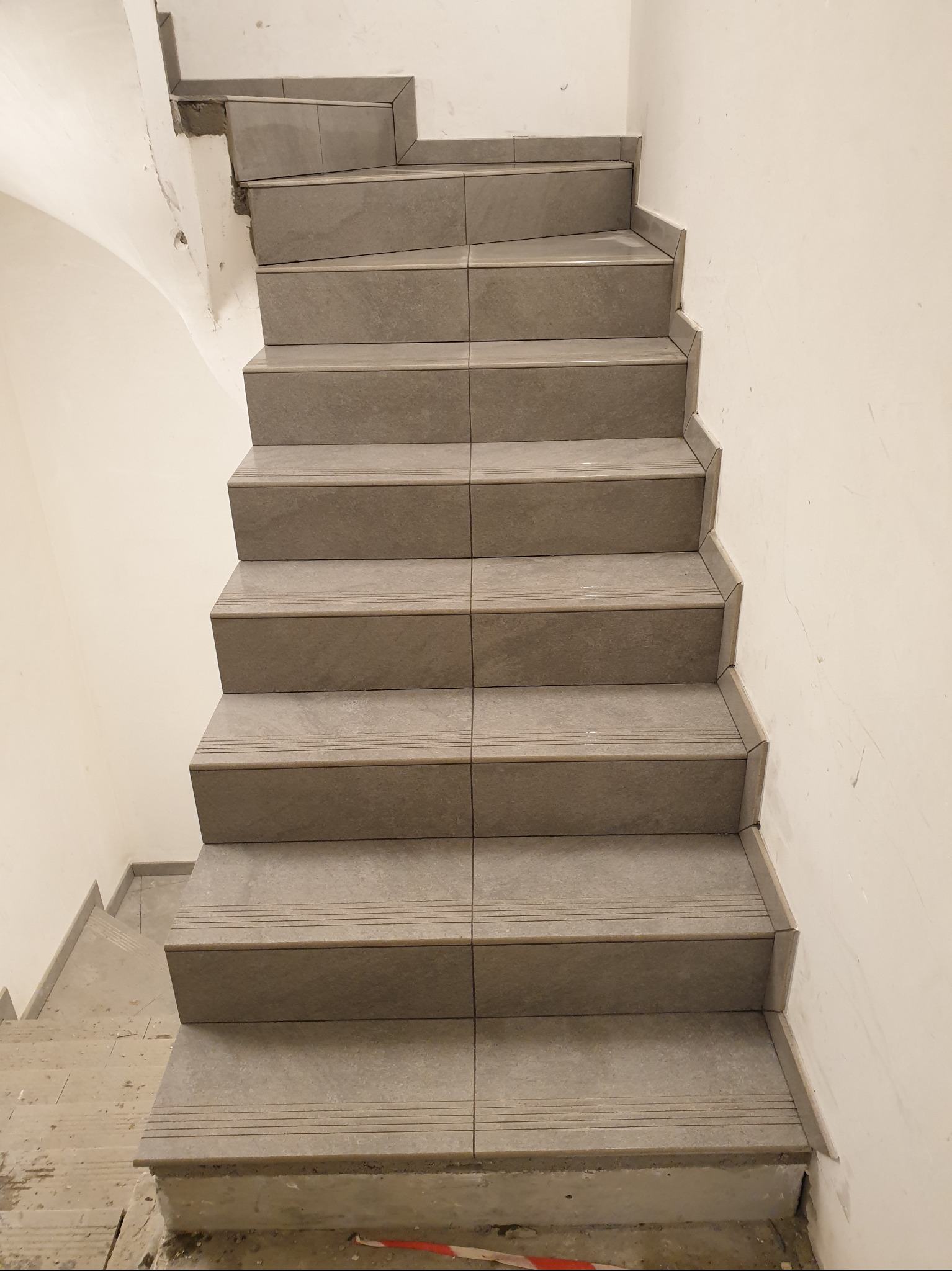 Wykończone schody z szarych płytek z delikatnym wzorem, widok z góry. Na stopniach widoczne subtelne, poziome rowki. Ściany pomalowane na jasny odcień bieli.