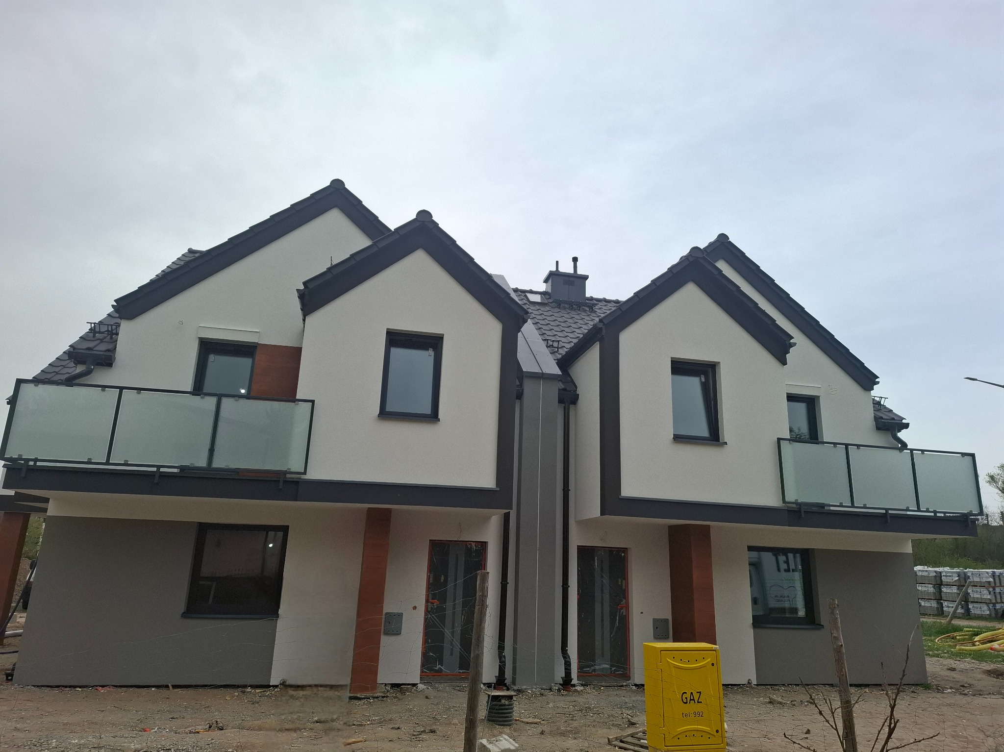 Dwa bliźniacze domy w nowoczesnym stylu z balkonami i szarą elewacją na dole. Widoczna skrzynka gazowa i materiały budowlane. Pochmurne niebo w tle.