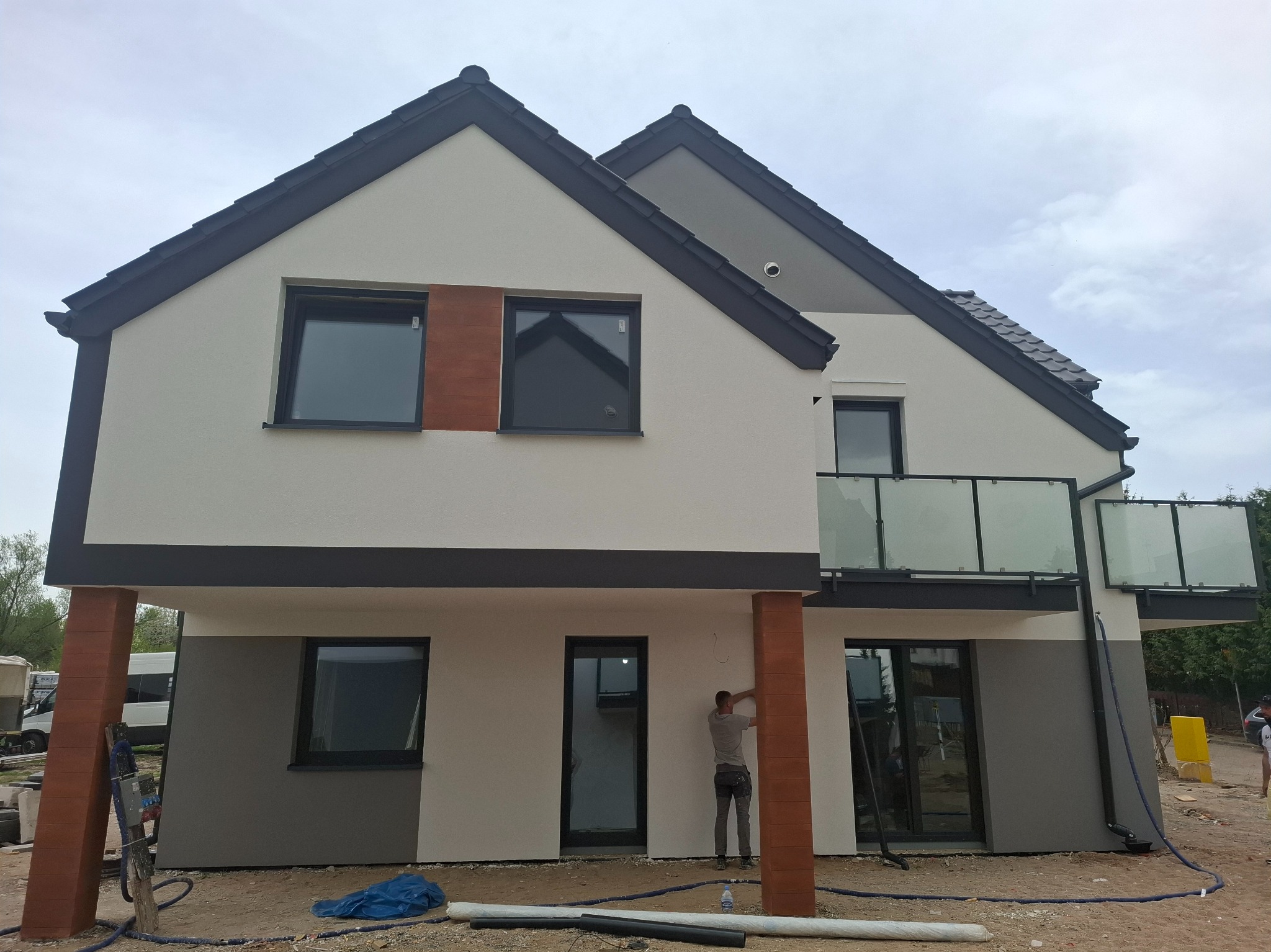 Dwukondygnacyjny dom z elewacją w odcieniach bieli i szarości, z elementami dekoracyjnymi imitującymi drewno. Pracownik wykonuje prace wykończeniowe przy kolumnie.