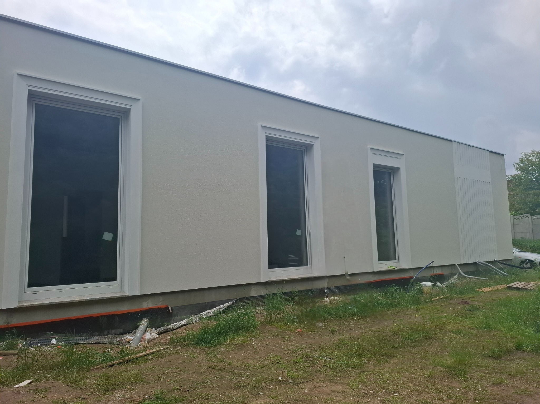Jasna elewacja budynku z trzema oknami o białych ramach, widok z zewnątrz, trawa i elementy instalacji przy fundamencie. Pochmurne niebo w tle.