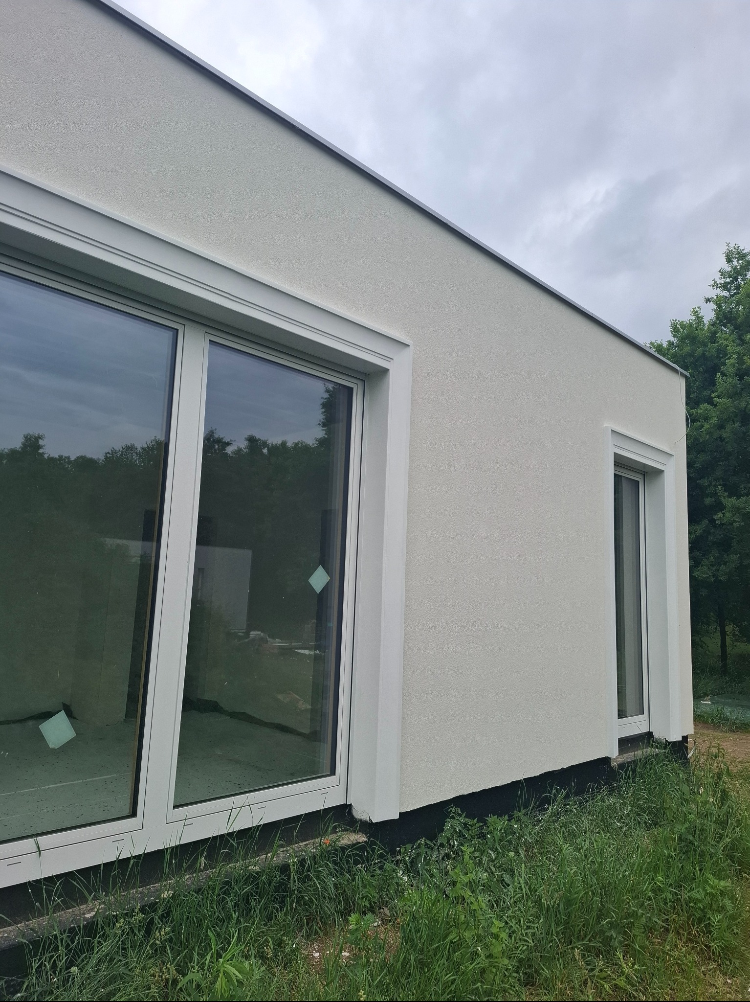 Boczny widok domu z jasną elewacją, dużymi oknami z białymi ramami i zielonym otoczeniem. Widoczna faktura tynku i detale wykończenia okien.