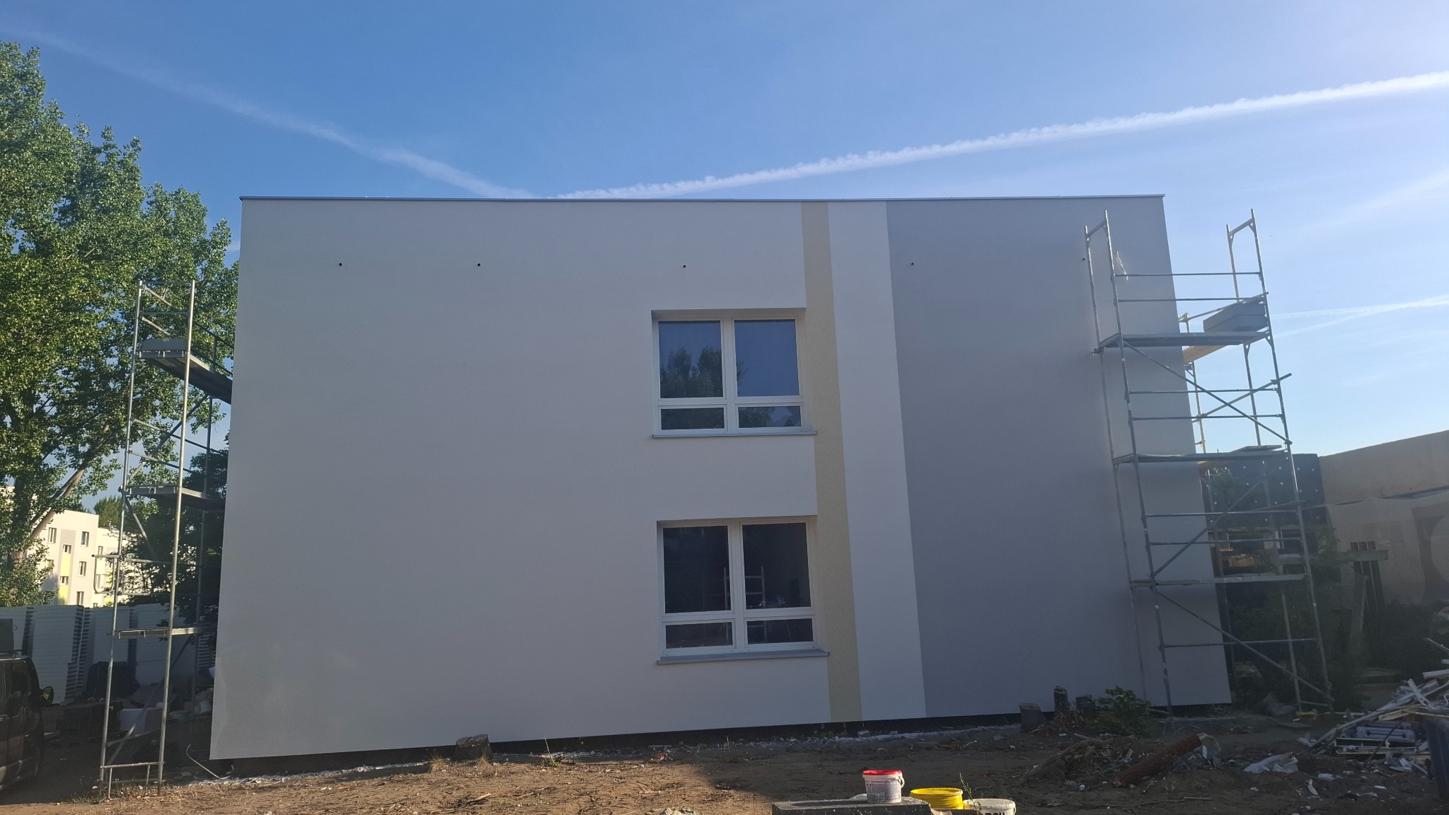 Dwukondygnacyjny budynek po malowaniu elewacji w odcieniach bieli i szarości, z widocznymi rusztowaniami i dwoma oknami na każdej kondygnacji.