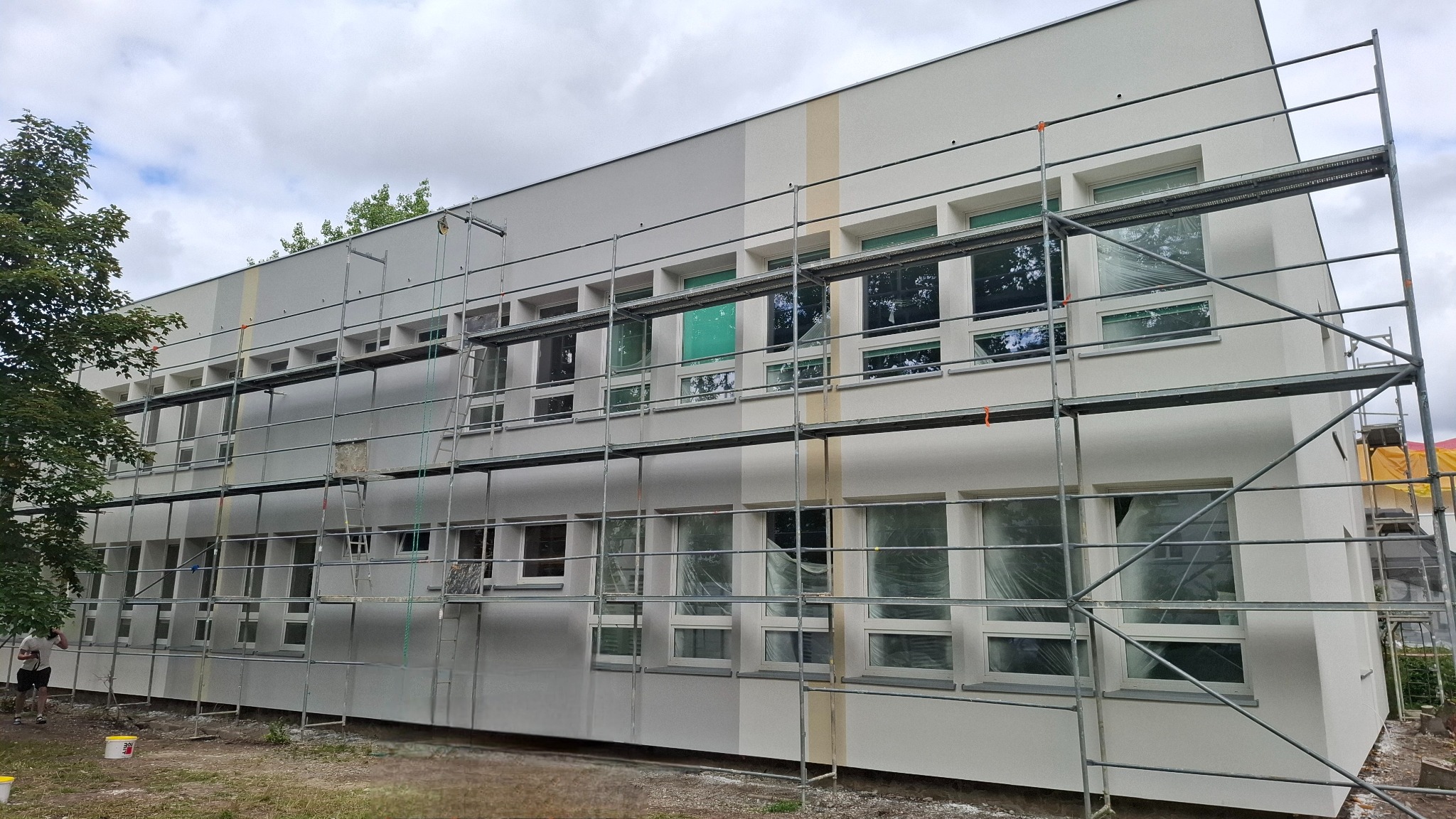 Budynek po renowacji elewacji, z widocznymi rusztowaniami i fragmentem zieleni. Jasna kolorystyka budynku z akcentami w odcieniach szarości i beżu.