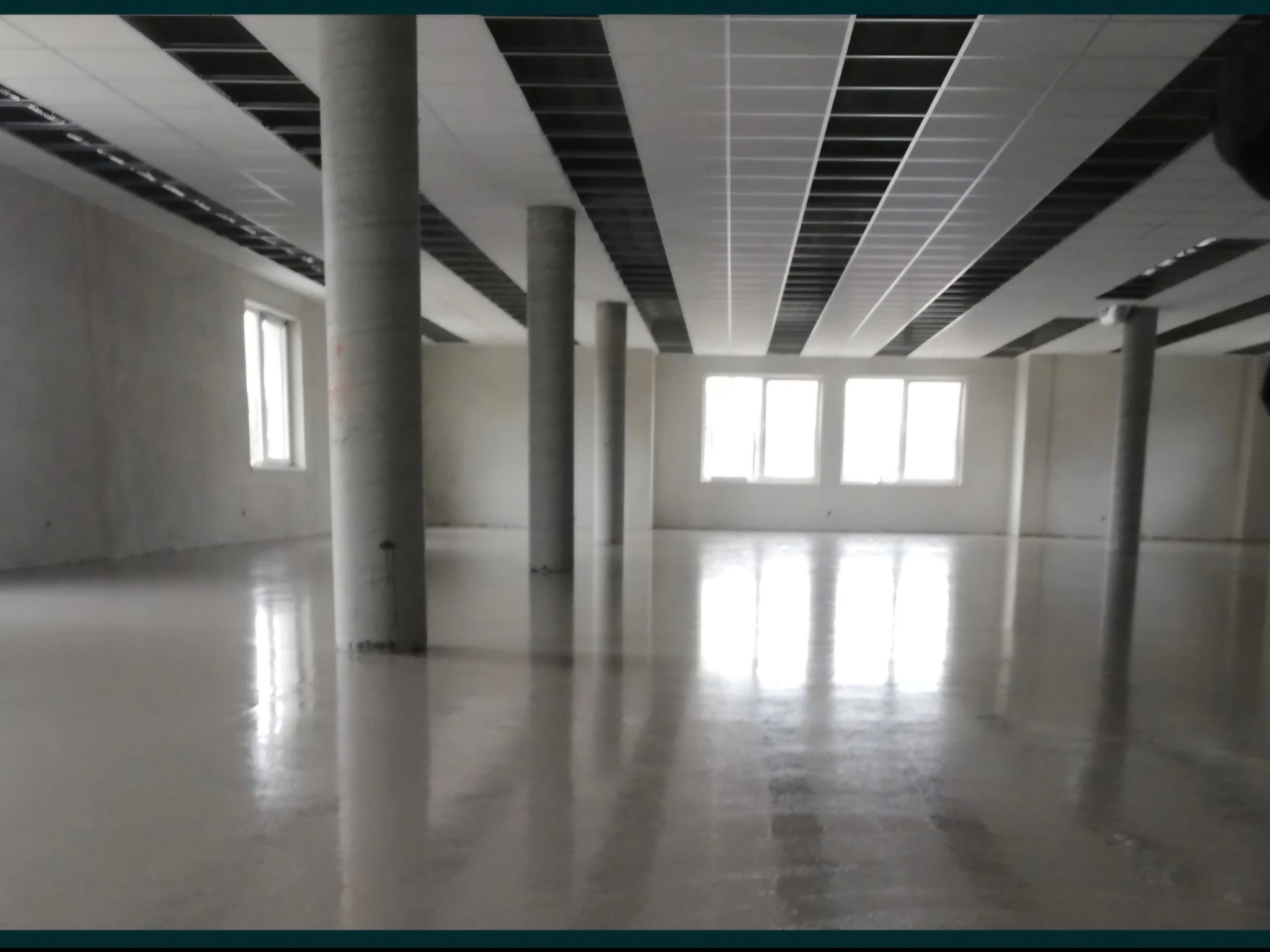 Wnętrze surowego biura z wylaną posadzką, widoczne kolumny, okna i częściowo zamontowany sufit podwieszany. Jasne światło dzienne.