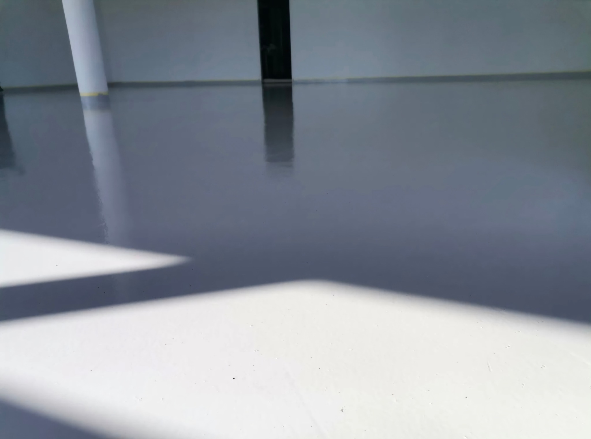 Gładka, szara posadzka przemysłowa z kolumną i drzwiami w tle, oświetlona naturalnym światłem tworzącym mocny cień w rogu. Nowoczesne wykończenie podłogi.