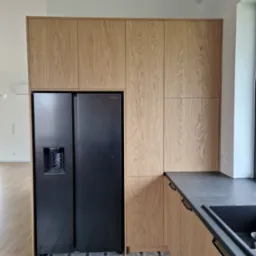 Jasna, drewniana zabudowa kuchenna z czarną lodówką side-by-side i ciemnym blatem. Na podłodze wzorzyste płytki. Minimalistyczny design, nowoczesne wykończenie.