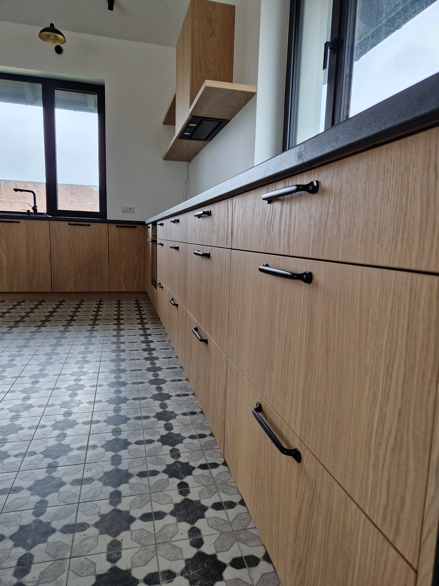 Nowoczesna kuchnia z drewnianymi frontami szafek i czarnymi uchwytami, wzorzysta podłoga. Widok na okno i blat. Minimalistyczny design.