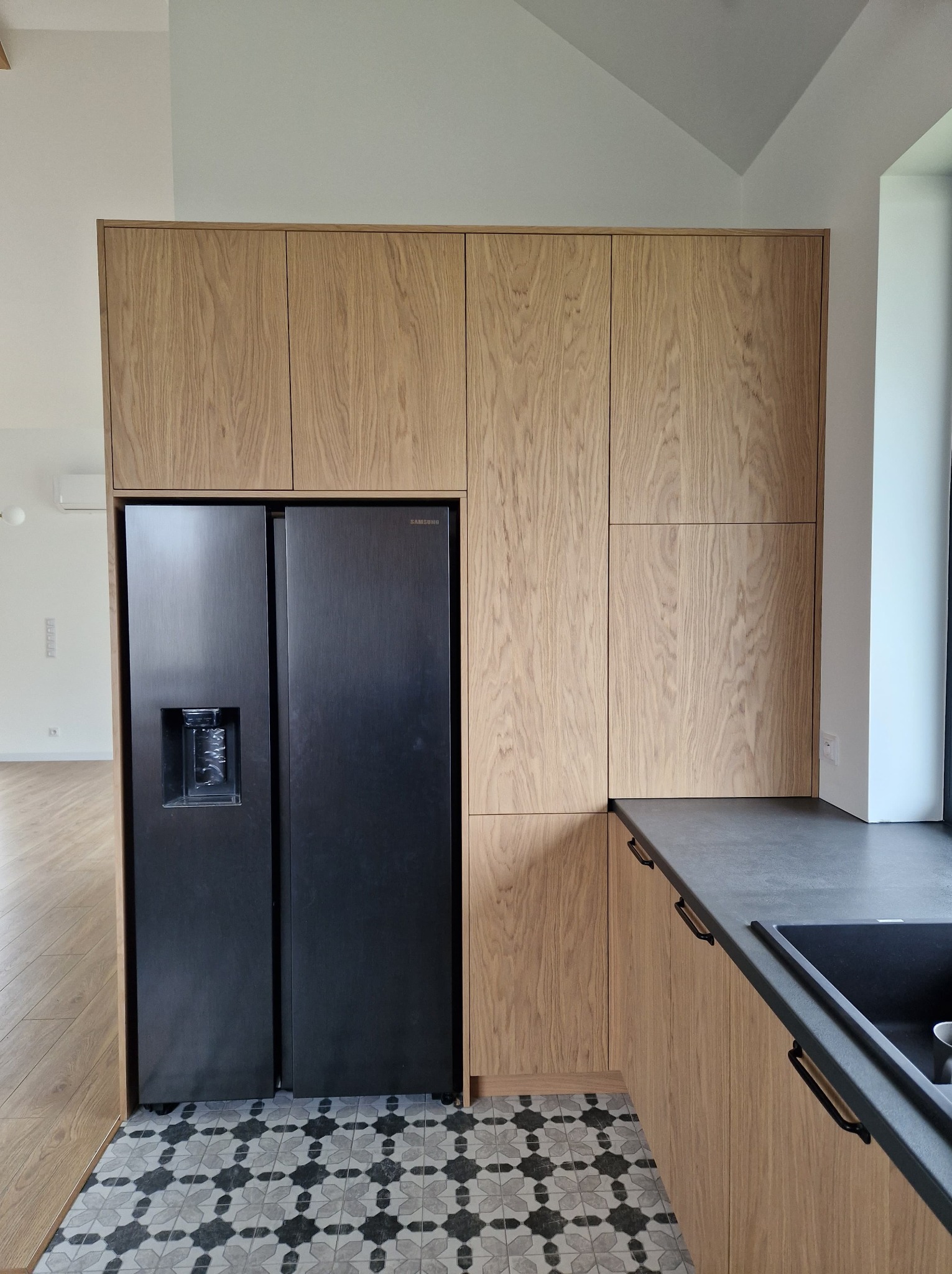 Jasna, drewniana zabudowa kuchenna z czarną lodówką side-by-side i ciemnym blatem. Na podłodze wzorzyste płytki. Minimalistyczny design, nowoczesne wykończenie.