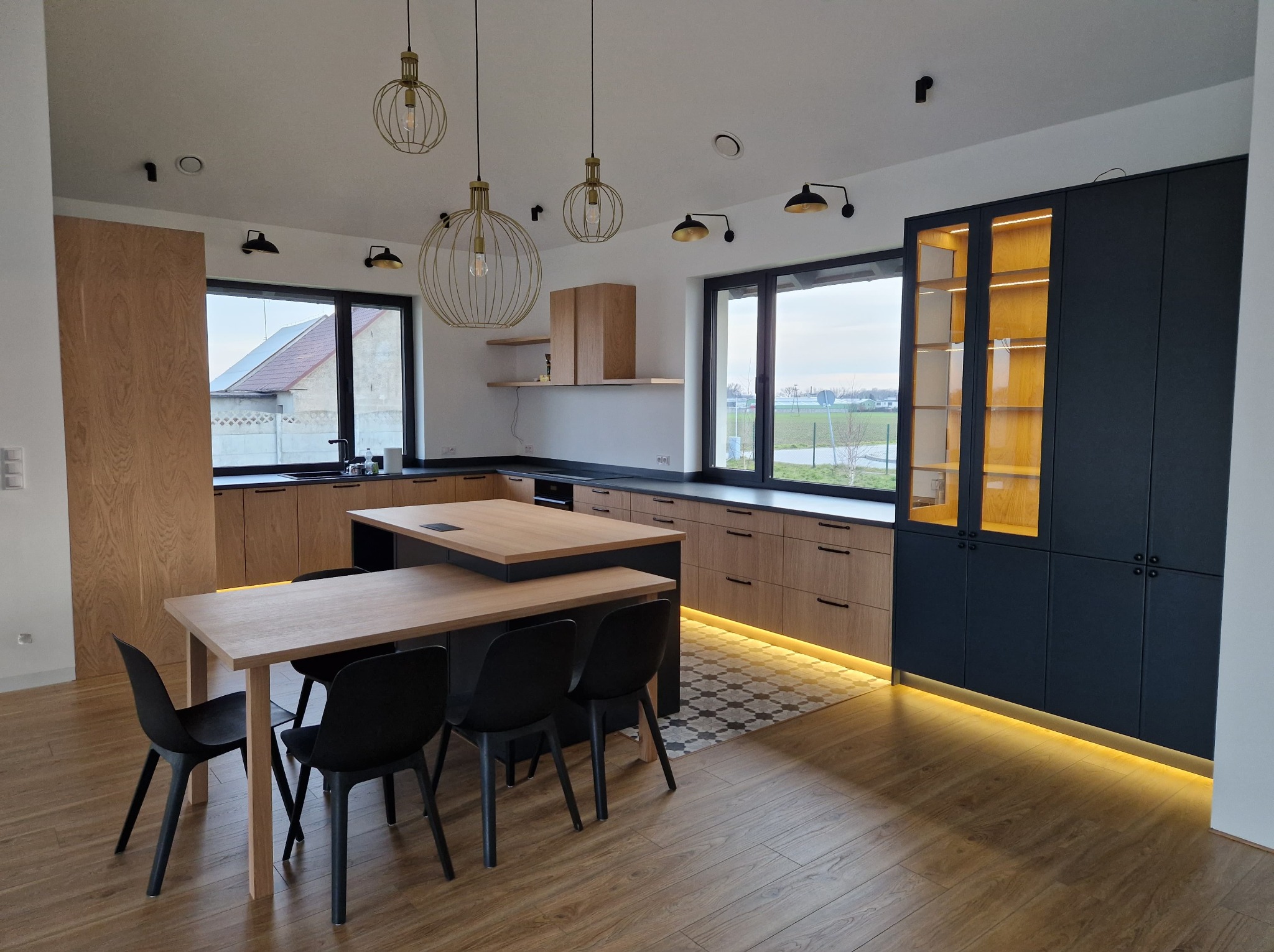 Nowoczesna kuchnia z drewnianymi frontami i wyspą, oświetlona wiszącymi lampami. Ciemne szafki z podświetleniem, stół z czarnymi krzesłami, podłoga drewniana.