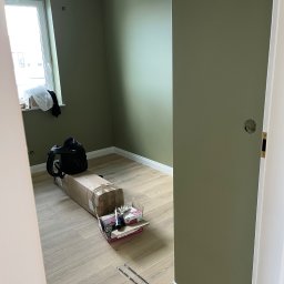 Denis Kareuka - Pokój po malowaniu na oliwkowy kolor, widoczne narzędzia i akcesoria malarskie, listwy przypodłogowe. Widok zza futryny drzwi.