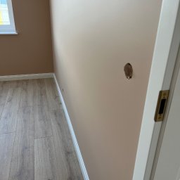 Denis Kareuka - Pokój przed remontem: dziura w beżowej ścianie, drewniana podłoga, biała listwa przypodłogowa i fragment białych drzwi. Widoczne okno z lewej strony.
