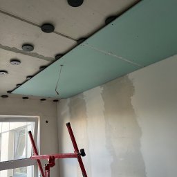 Denis Kareuka - Montaż płyty gipsowej na suficie za pomocą podnośnika. Widoczne elementy mocujące i częściowo wyszpachlowana ściana. W tle okno z widokiem na budynek.