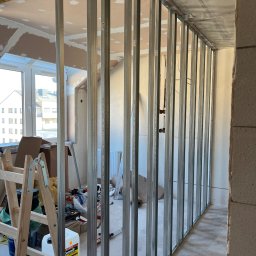 Denis Kareuka - Metalowa konstrukcja ścianki działowej na tle okna i surowego wnętrza. Widoczne narzędzia i materiały budowlane, w tym drabina i pojemnik z gruntem Ceresit.