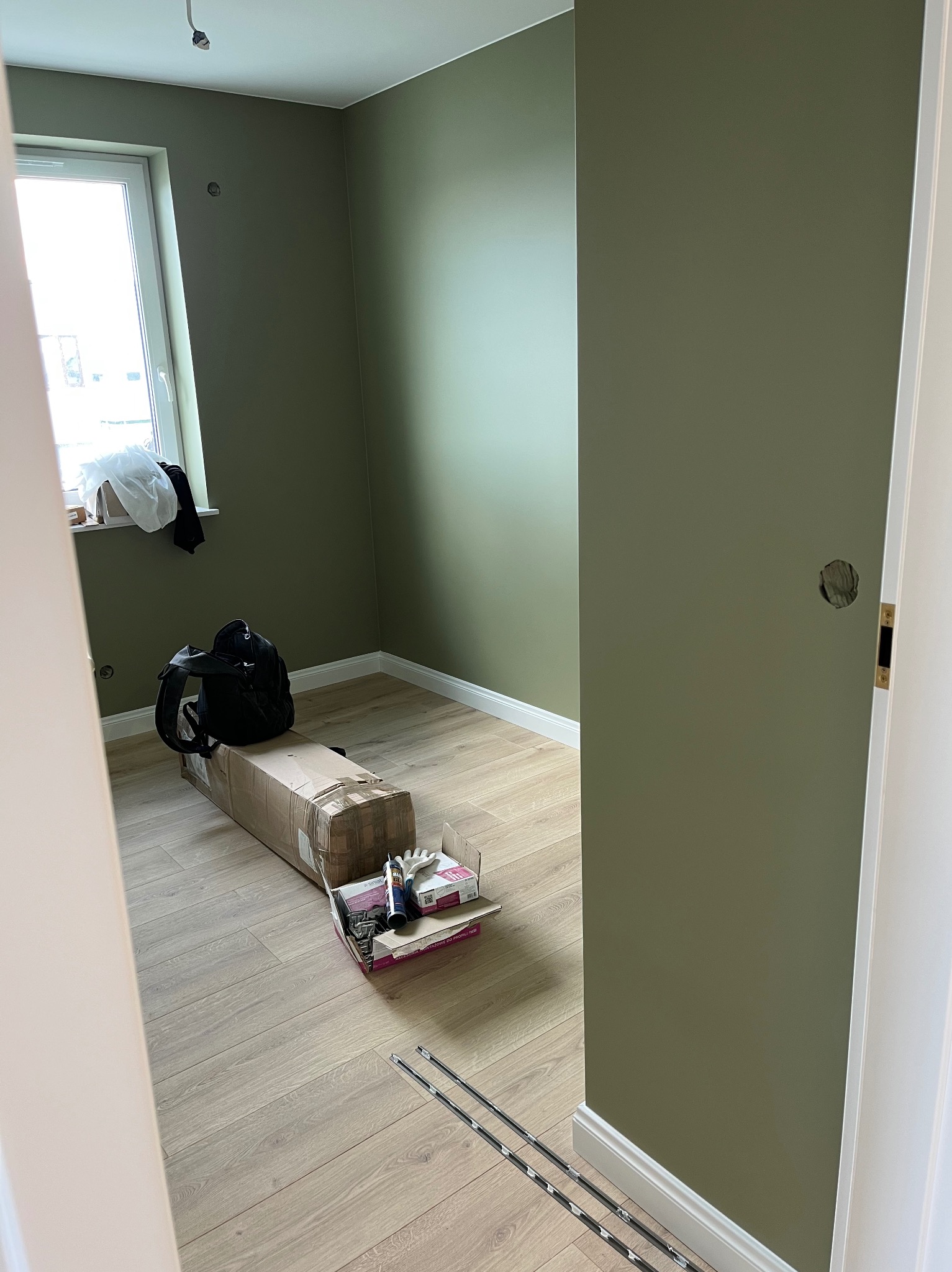 Pokój po malowaniu na oliwkowy kolor, widoczne narzędzia i akcesoria malarskie, listwy przypodłogowe. Widok zza futryny drzwi.