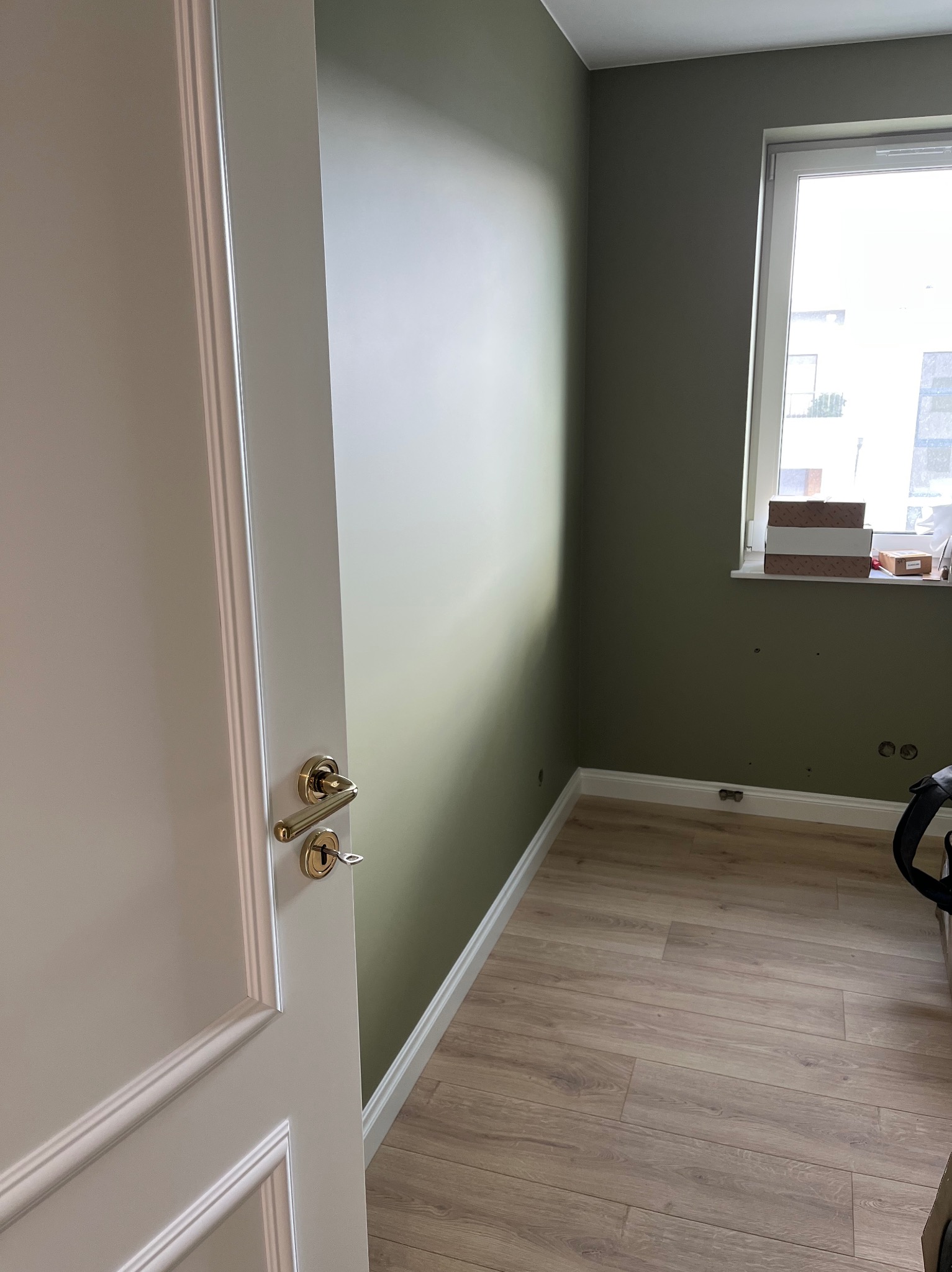 Pomalowane na oliwkowo ściany pokoju z drewnianą podłogą, widok zza uchylonych, białych drzwi z ozdobnymi listwami i złotą klamką. Na parapecie kartony.