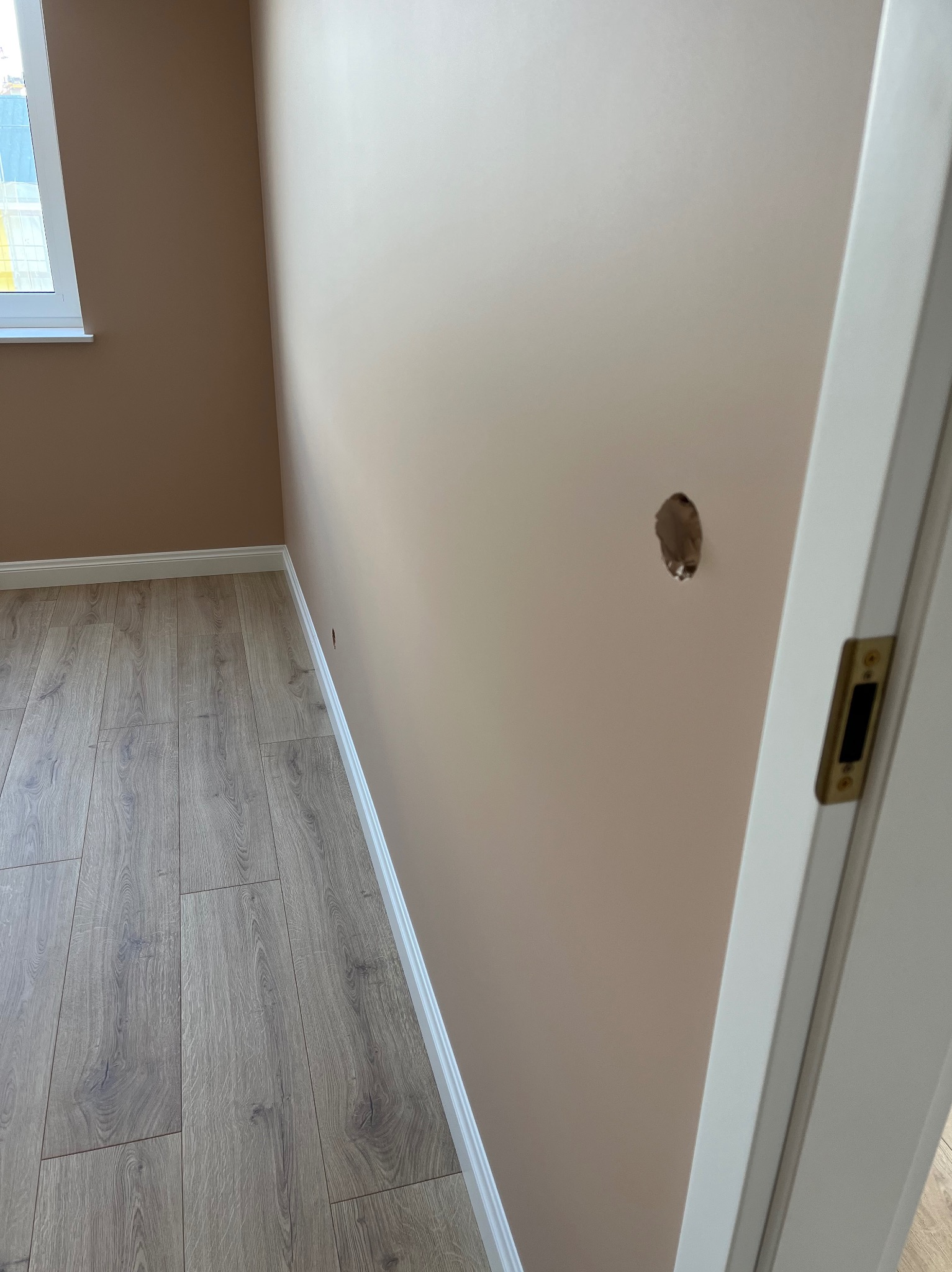 Pokój przed remontem: dziura w beżowej ścianie, drewniana podłoga, biała listwa przypodłogowa i fragment białych drzwi. Widoczne okno z lewej strony.
