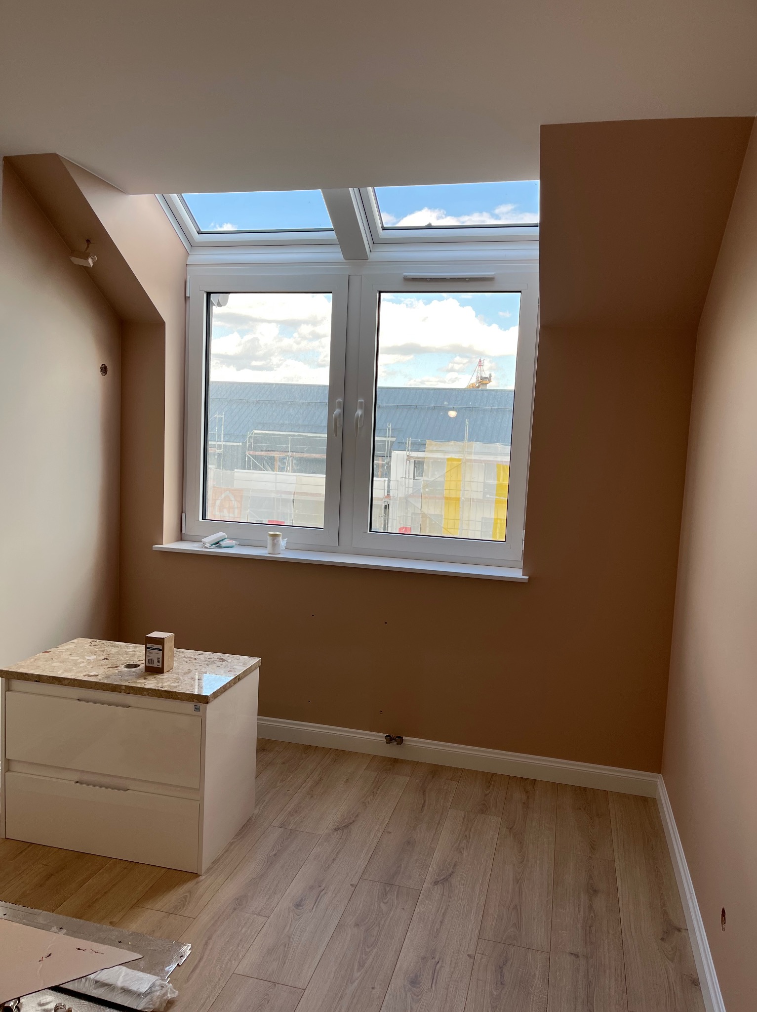 Pokój na poddaszu po malowaniu ścian na beżowo, z oknem dachowym i komodą z blatem imitującym lastryko. Widoczne listwy przypodłogowe i panele podłogowe.