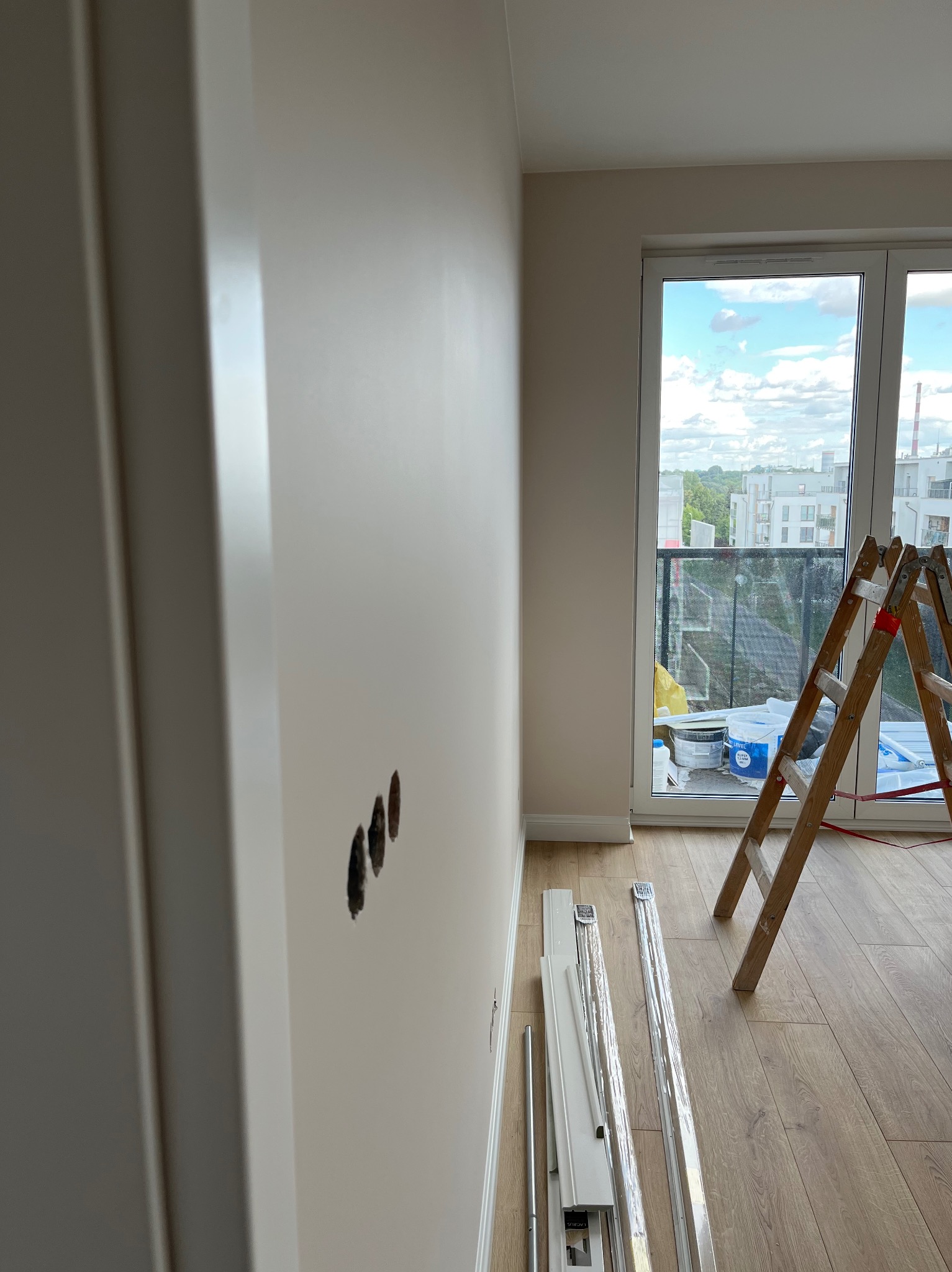 Ściana po szpachlowaniu z otworami na gniazdka, drabina i listwy przypodłogowe przy oknie balkonowym. Widok na miasto w tle. Jasne wnętrze w trakcie prac.