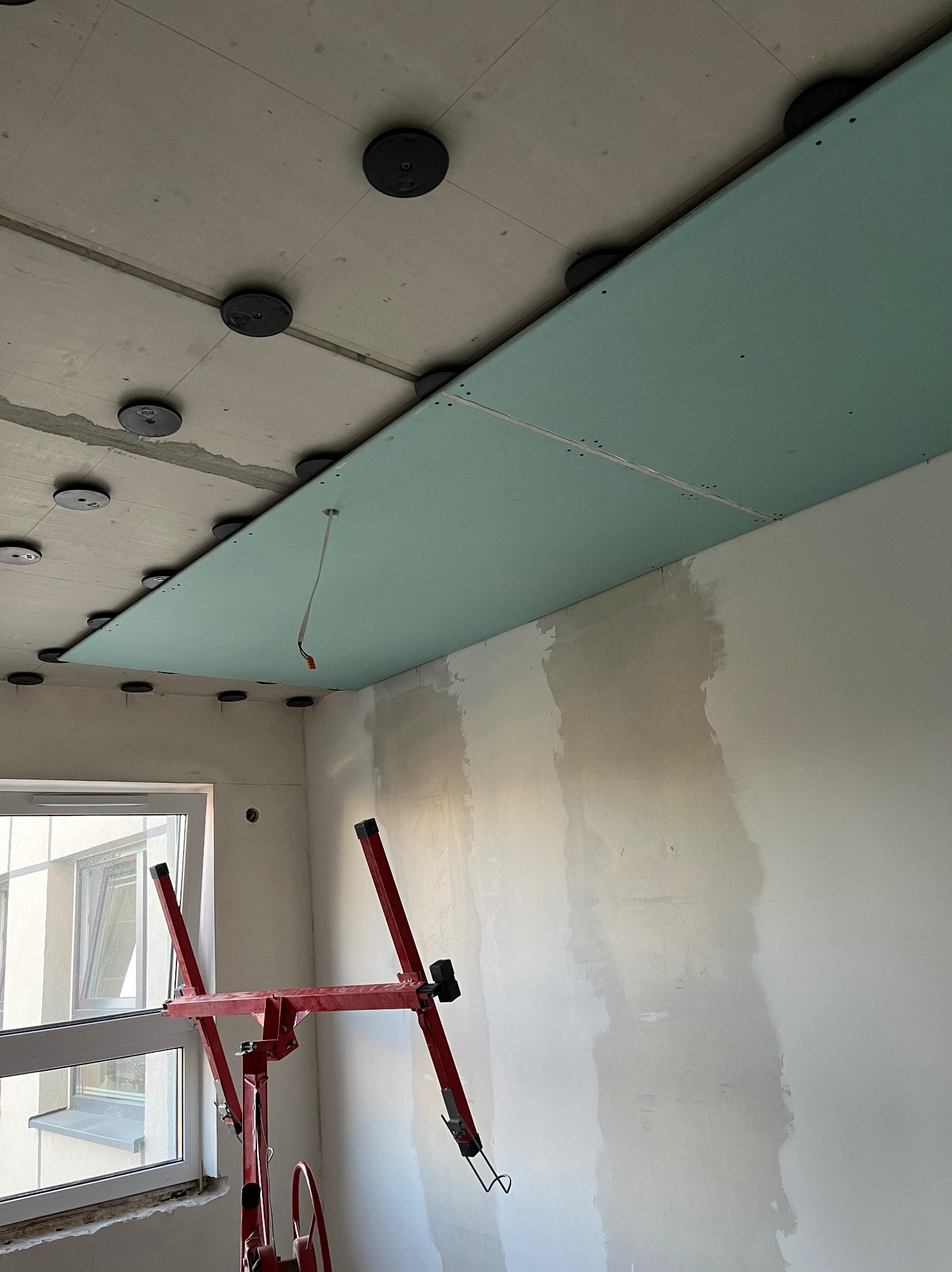 Montaż płyty gipsowej na suficie za pomocą podnośnika. Widoczne elementy mocujące i częściowo wyszpachlowana ściana. W tle okno z widokiem na budynek.