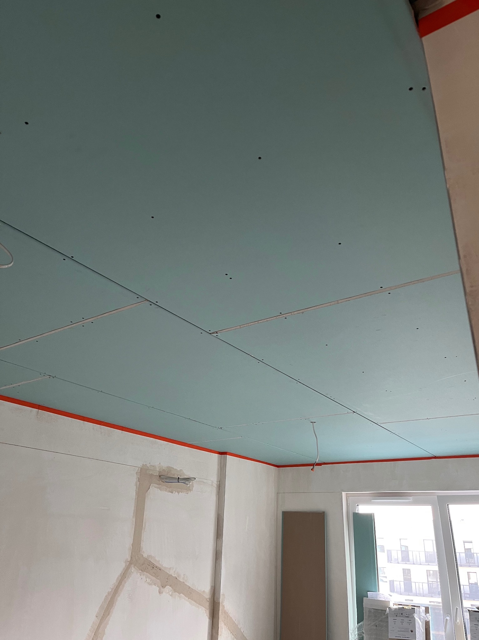 Niebieskie płyty gipsowo-kartonowe zamontowane na suficie, widoczne łączenia i ślady po wkrętach. Ściany częściowo wyszpachlowane, okno balkonowe w tle.