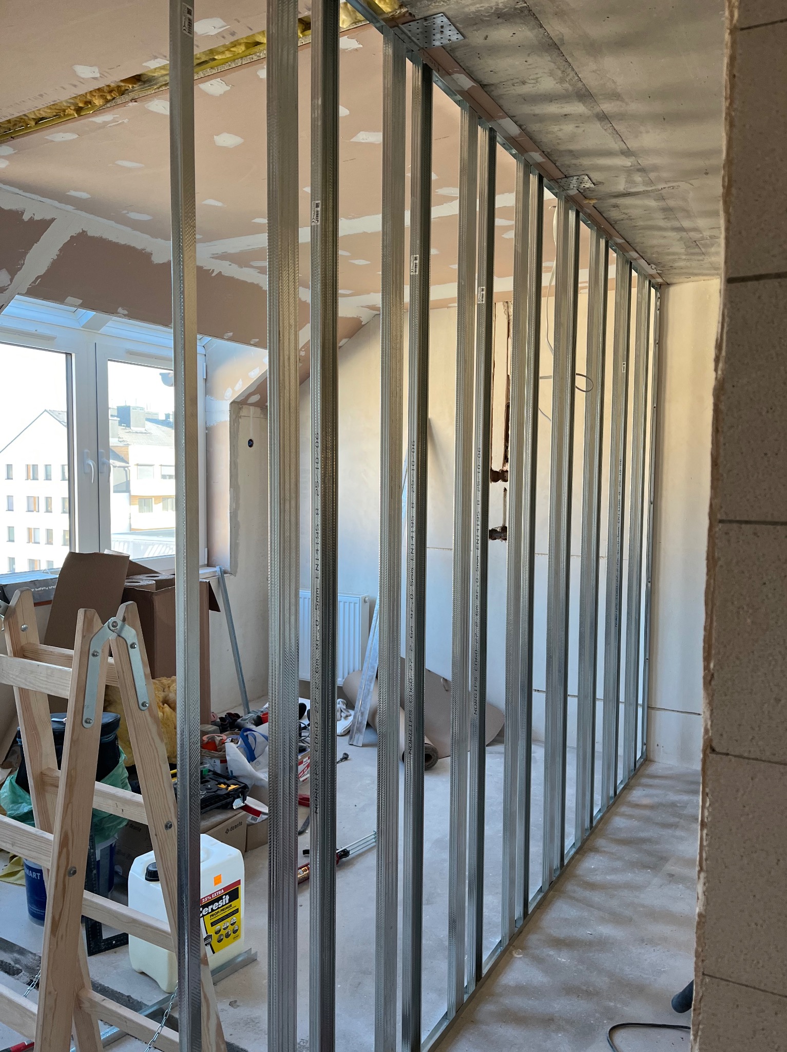 Metalowa konstrukcja ścianki działowej na tle okna i surowego wnętrza. Widoczne narzędzia i materiały budowlane, w tym drabina i pojemnik z gruntem Ceresit.