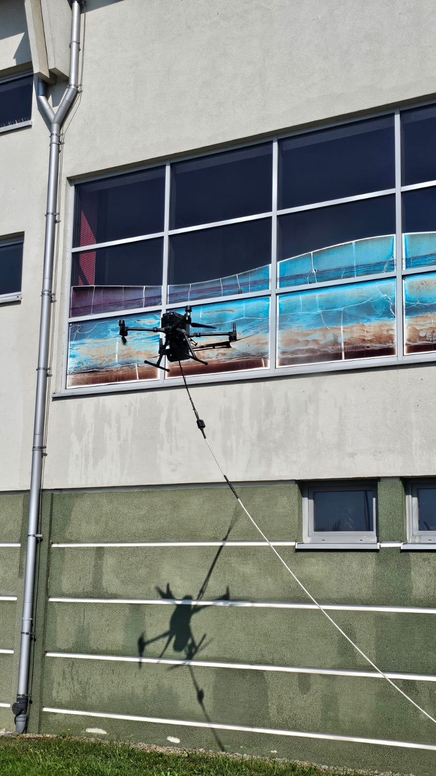 Dron z lancą teleskopową czyści okna na wysokości. Widoczny cień drona na elewacji budynku. Nietypowe rozwiązanie do mycia okien.