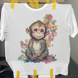 iWallDesign Kamil Musioł - Biała koszulka z nadrukiem uroczego, siedzącego małpki w otoczeniu akwarelowych kwiatów, przymocowana taśmą do podłoża, prezentacja nadruku na odzieży.