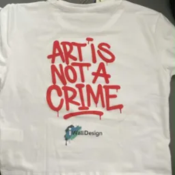 Biała koszulka z czerwonym napisem 'Art is not a crime' i logo 'iWallDesign', leżąca na szarym tle. Widoczna metka. Nietypowa czcionka i układ.