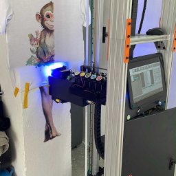 iWallDesign Kamil Musioł - Biała koszulka z nadrukiem małpki, przymocowana do ściany taśmą. Drukarka do nadruków na odzieży podczas pracy, widoczne kartridże z kolorami i niebieskie światło.