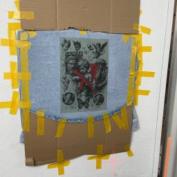 iWallDesign Kamil Musioł - Jasna kurtka dżinsowa z nadrukiem aniołów i postaci Herculesa, przymocowana taśmą do ściany, przygotowana do dalszej obróbki przy użyciu lasera.