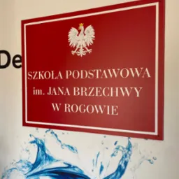 Czerwona tablica informacyjna z godłem Polski i napisem 'Szkoła Podstawowa im. Jana Brzechwy w Rogowie', umieszczona na ścianie z niebieskim motywem wodnym.
