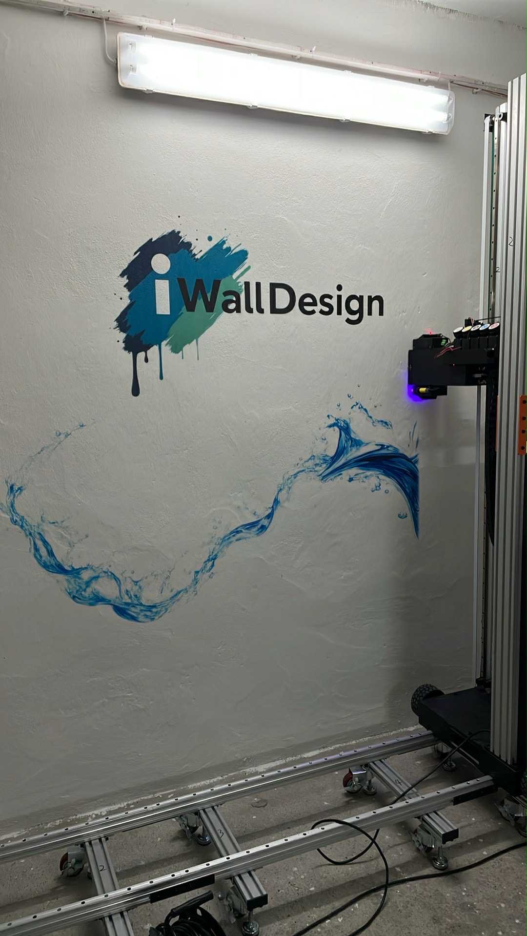 Drukarka ścienna w trakcie nanoszenia niebieskiego wzoru fal na szarą ścianę z fakturą; widoczny logo 'iWallDesign' i oświetlenie górne.